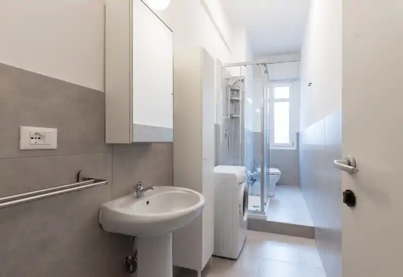 Bright, modern bathroom with a pedestal sink, mirrored cabinet, shower enclosure, toilet and a washing machine in a clean, neutral tile setting.
