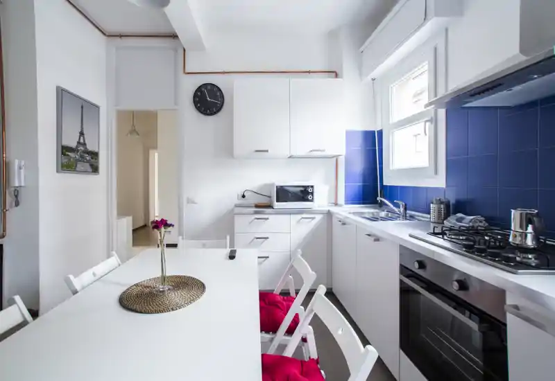 Cucina moderna e luminosa con mobili bianchi, paraschizzi piastrellato blu, forno a incasso e piano cottura a gas, con tavolo e sedie: ambiente pulito e funzionale ideale per cucinare e pranzi informali.