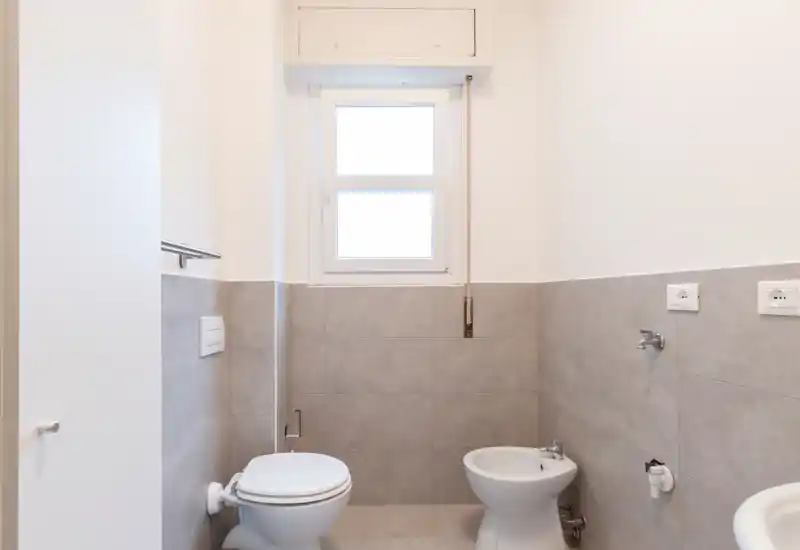 Clean, minimal bathroom with a toilet, bidet and tiled half-wall beneath a bright window; neutral gray tiles and white fixtures create a fresh, modern look.