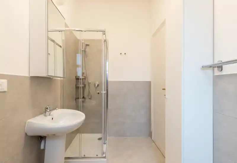 Clean modern bathroom with a corner glass shower, wall-mounted mirror cabinet and pedestal sink. Neutral gray tiles and bright white walls create a minimalist, easy-to-maintain look.