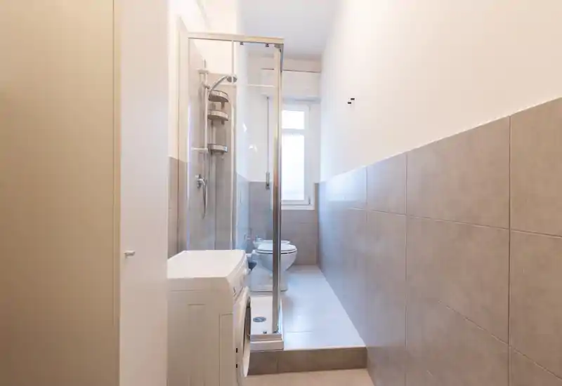 Narrow modern bathroom with a glass-enclosed shower, toilet and neutral gray wall tiles; bright natural light from a window creates a clean, minimalist feel.