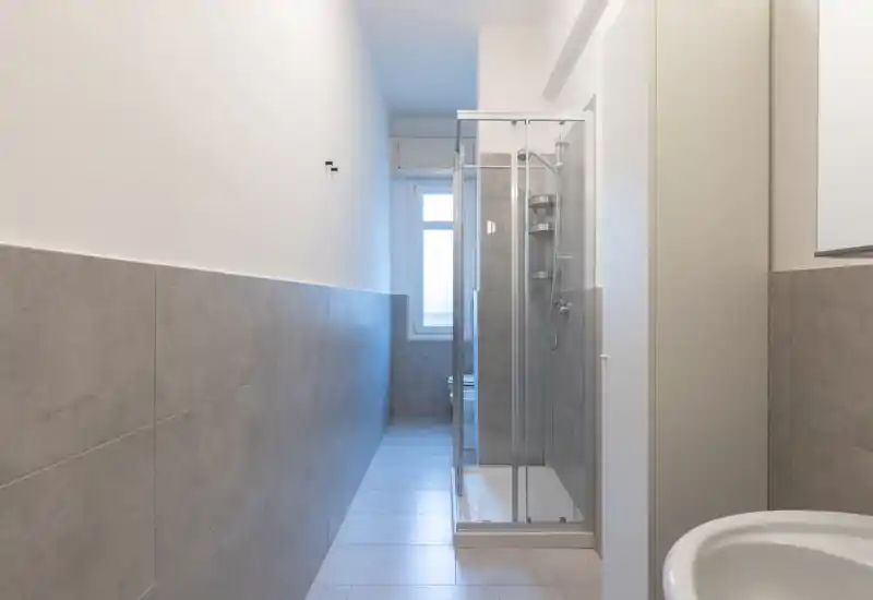 Bright, modern narrow bathroom with a glass shower enclosure, grey wall tiles and a white sink visible in the foreground.