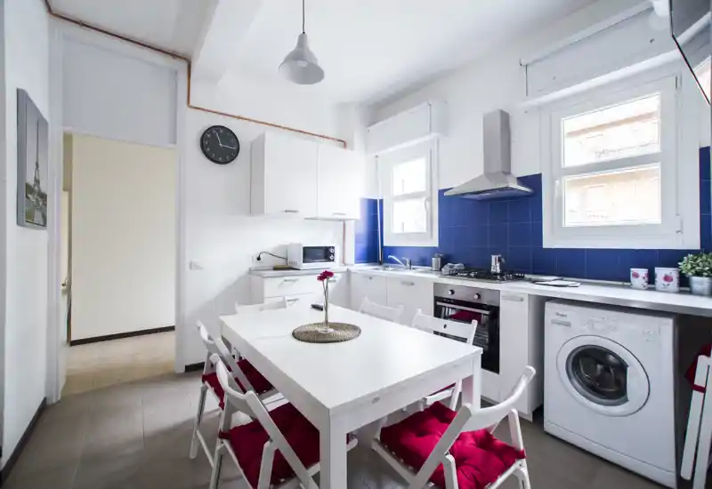 Cucina moderna e luminosa con tavolo da pranzo integrato. Pensili bianchi, paraschizzi piastrellato blu intenso, piano cottura a gas e lavatrice creano uno spazio pratico e di stile per cucinare e mangiare.