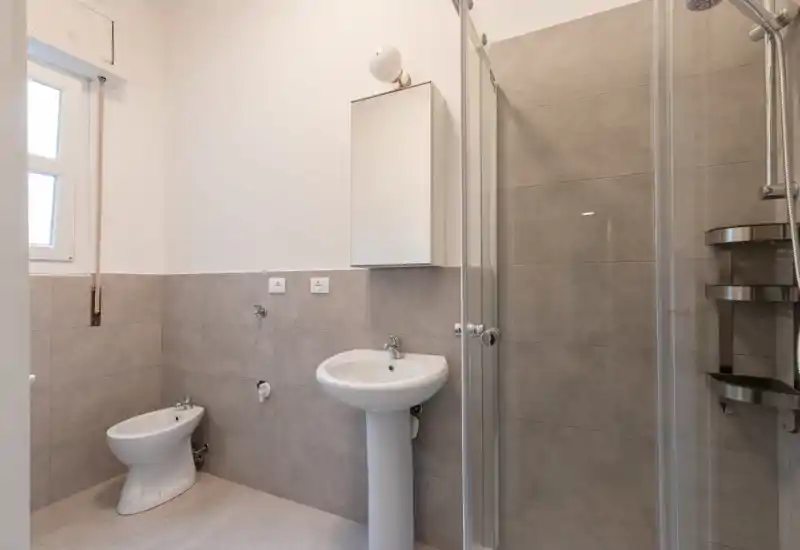 Clean, modern bathroom with pedestal sink, bidet, and a glass-enclosed shower; neutral grey tiles and a wall cabinet provide practical storage.