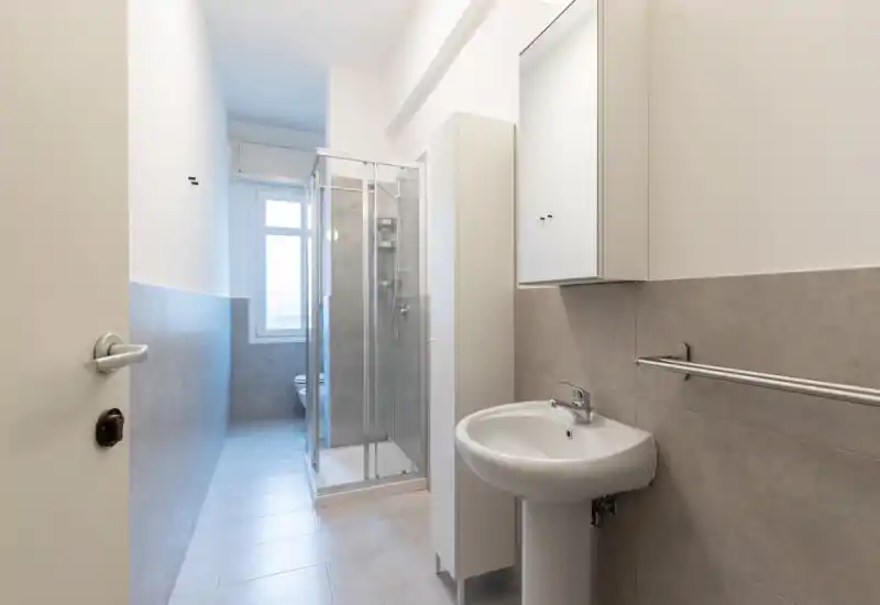 Modern, compact bathroom with a pedestal sink, mirrored cabinet, towel rail and a glass-enclosed shower. Neutral tiles and natural light from a window create a clean, functional space ideal for short-term rentals.