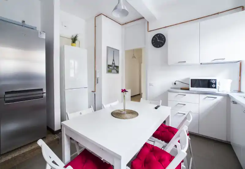 Bright modern kitchen with a white dining table, red seat cushions and stainless appliances. Clean lines and natural light make it ideal for casual meals.