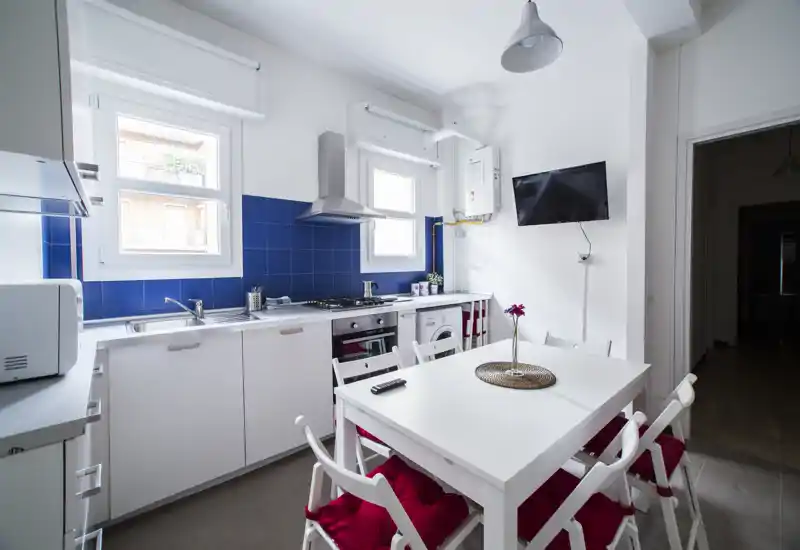 Cucina moderna e luminosa con mobili bianchi, piano cottura a gas e tavolo da pranzo con cuscini rossi — ben illuminata e ordinata.