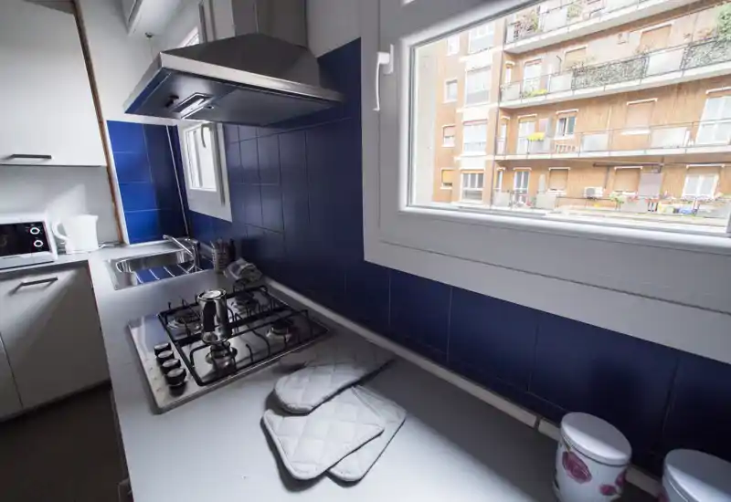 Cocina moderna y compacta con placa de gas, fregadero de acero inoxidable y salpicadero de azulejos azules; una gran ventana aporta luz natural y vistas a la calle.