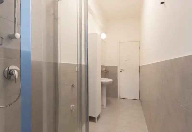 Modern narrow bathroom with a glass shower, pedestal sink, tall storage cabinet and neutral grey tiles, well lit and minimal.