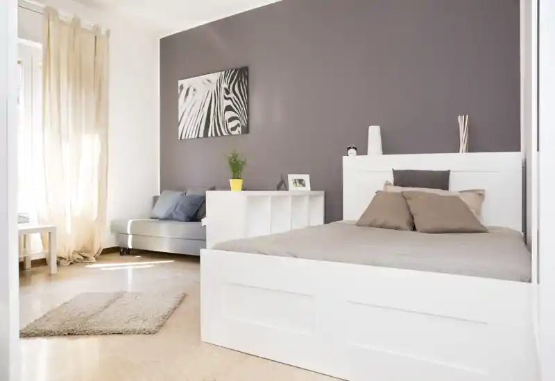 Bright, modern bedroom with a large white bed in the foreground, neutral linens, a small sofa by the window and minimalist decor.