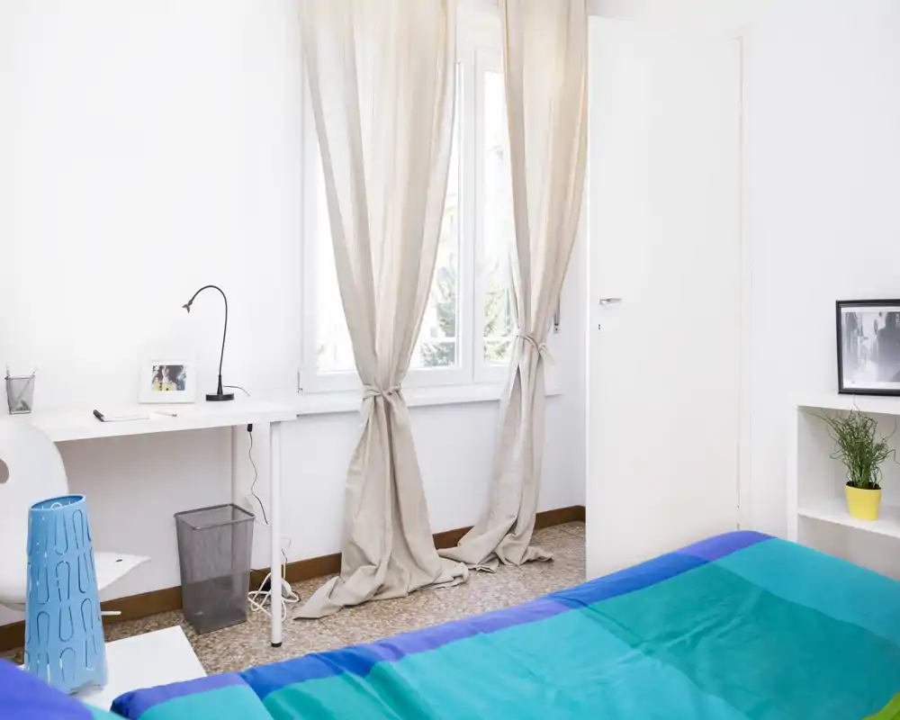 Camera da letto luminosa e minimal con una copriletto colorata, ampia finestra con tende chiare e una scrivania semplice — ideale per un alloggio accogliente e ben illuminato.