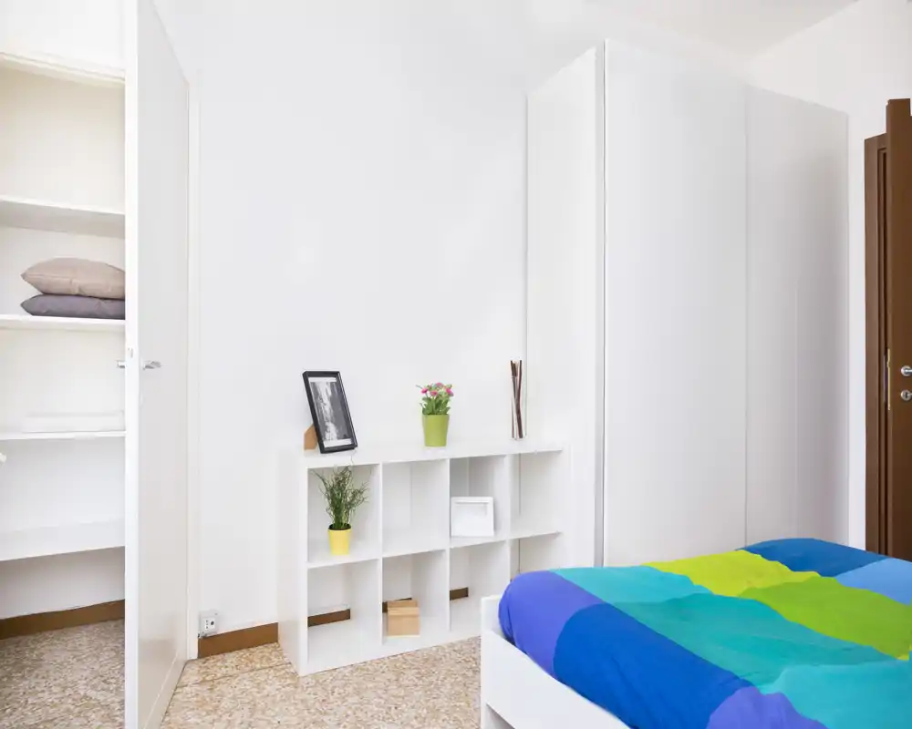 Camera da letto luminosa e minimal con una trapunta colorata, scaffalatura bianca e armadio; stile pulito e moderno con buona luce naturale.