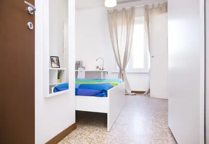 Camera singola luminosa e minimale con letto bianco, biancheria colorata, scrivania e luce naturale dalla finestra — ordinata e pronta per gli ospiti.