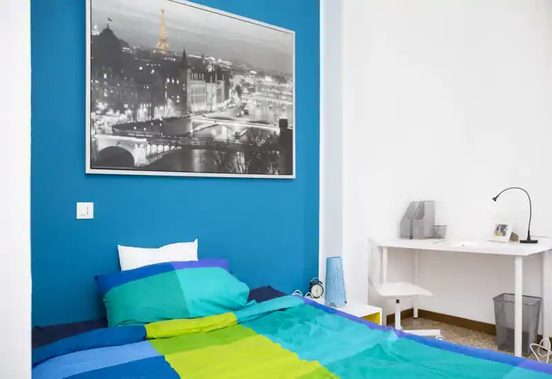 Camera da letto luminosa e moderna con biancheria colorata, una grande stampa della città sopra il letto e una piccola postazione di lavoro laterale. L'ambiente è arioso e fresco con una parete accentuata blu e una scrivania funzionale.