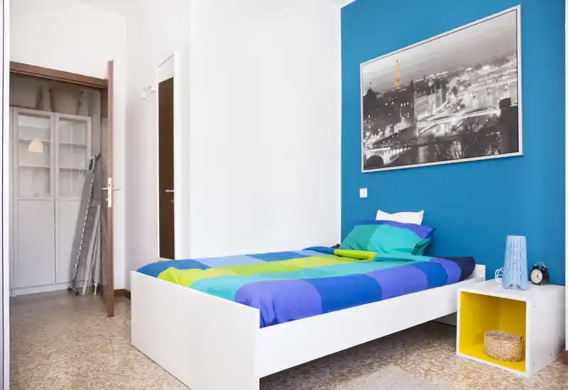 Camera singola luminosa e ben arredata con letto dai toni blu e verdi, comodino a cubo, quadro a parete e capiente armadio. Ideale per affitti brevi o studenti.