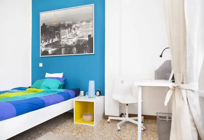 Camera da letto moderna e luminosa con un letto singolo colorato, un cubo portaoggetti come comodino e una piccola scrivania vicino alla finestra — pulita e di stile.