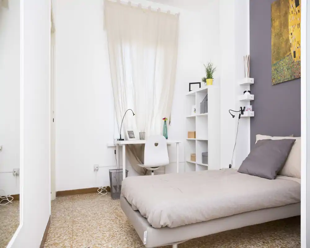 Camera da letto singola luminosa e minimal con letto ordinato, scrivania compatta vicino alla finestra e mensole aperte — ideale per uno studente o occupante singolo.