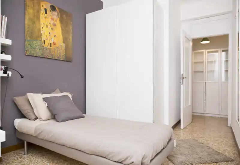 Camera da letto singola luminosa e ben allestita con letto ordinato, cuscini morbidi e grande armadio bianco; un quadro di buon gusto e toni caldi creano un’atmosfera accogliente.