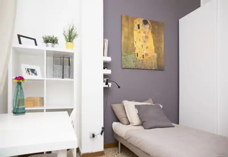 Camera da letto compatta e luminosa con un letto singolo con biancheria neutra, cuscini decorativi e un quadro sopra. Una libreria bianca e una scrivania creano un angolo funzionale e accogliente, ideale per una persona.