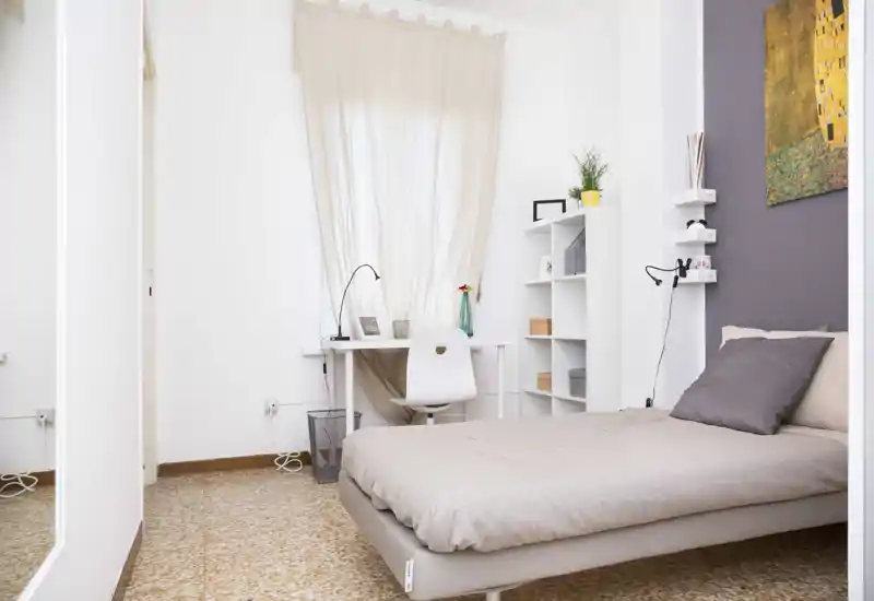 Camera da letto singola luminosa e minimal con letto ordinato, scrivania compatta vicino alla finestra e mensole aperte — ideale per uno studente o occupante singolo.