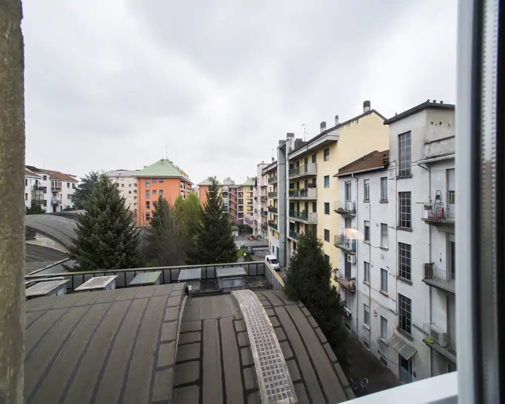 Vista esterna urbana da una finestra con edifici residenziali vicini, cime di alberi e tetti sotto un cielo nuvoloso — adatta per mostrare il contesto e i dintorni dell'immobile.