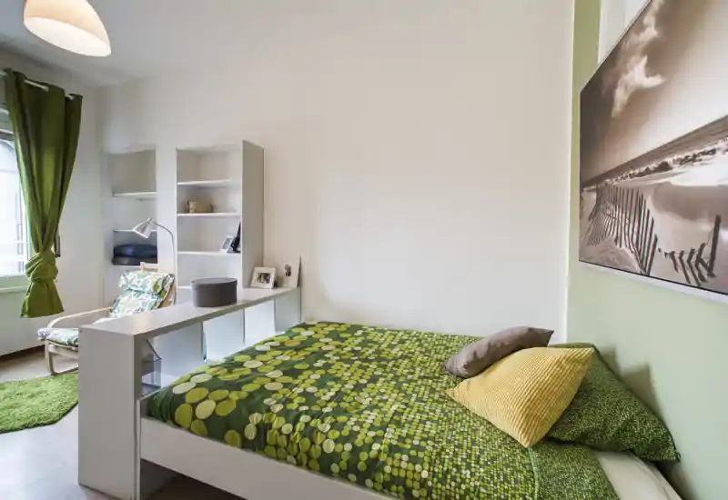 Camera da letto luminosa e ben allestita con un letto matrimoniale verde a motivi, cuscini accoglienti e arredi coordinati, poltrona per la lettura e scaffalatura vicino alla finestra.