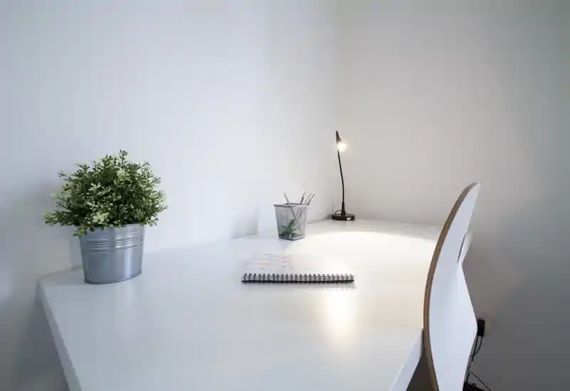 Spazio di lavoro minimalista e luminoso con scrivania bianca, piccolo vaso con pianta, un taccuino e una lampada da tavolo accesa — ordinato e ideale per concentrarsi.