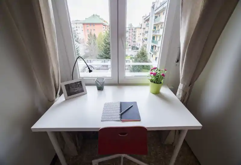 Bright workspace by a large window with a simple white desk, red chair, notebook and a small potted plant—ideal for a compact home office with pleasant city views.
