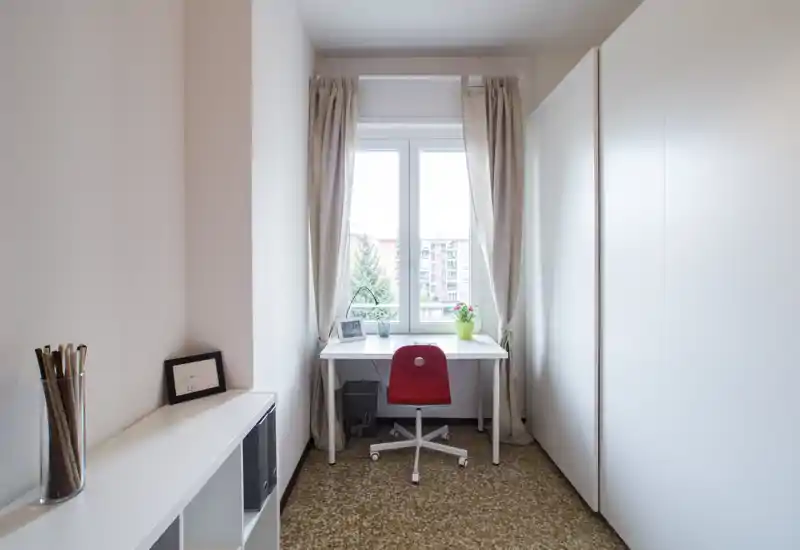 Bright minimalist small room set up as a home office nook with a white desk by the window, red swivel chair, wardrobe on the right and shelving on the left.