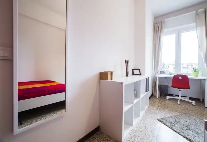 Small, bright bedroom seen mainly through a wall mirror: a fitted shelving unit and a desk by the window with a red chair. Natural light fills the space, which is simply furnished and tidy.