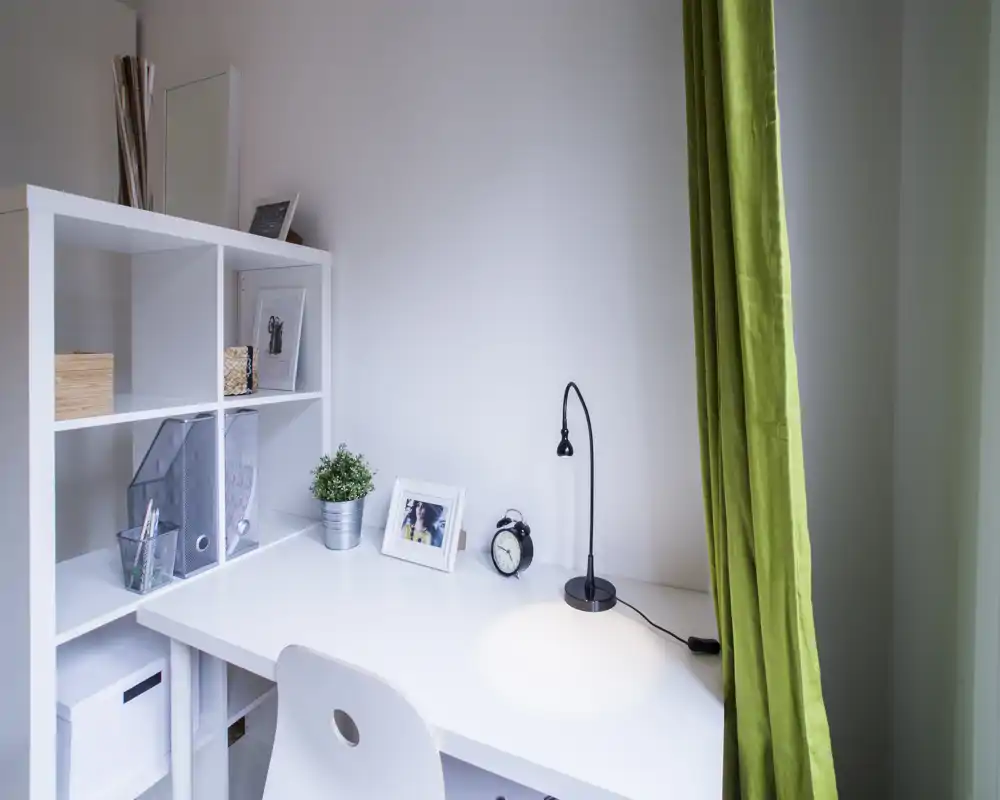Espace de travail à domicile lumineux et minimaliste avec bureau blanc, étagère, petite lampe, photo encadrée et rideau vert — parfait pour mettre en valeur un coin bureau soigné dans une annonce.