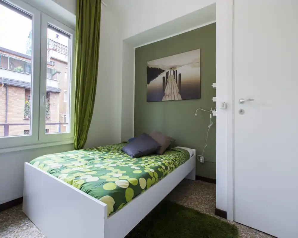 Petite chambre aménagée avec un lit simple à couette verte à motifs, deux oreillers, une œuvre accrochée au-dessus de la tête de lit et une grande fenêtre apportant la lumière naturelle.