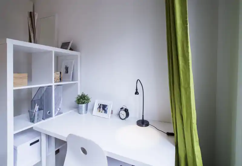 Bright, minimal home workspace with a white desk, shelving unit, small lamp, framed photo and a green curtain — ideal for a tidy rental listing highlighting a study nook.