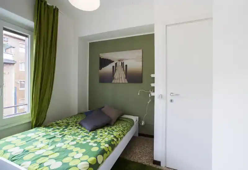 Chambre simple lumineuse avec un lit recouvert d'une couette verte à motifs, des rideaux verts assortis et un tableau paysager au-dessus de la tête de lit. La lumière naturelle entrant par la fenêtre donne une atmosphère fraîche, propre et accueillante, idéale pour une annonce locative.