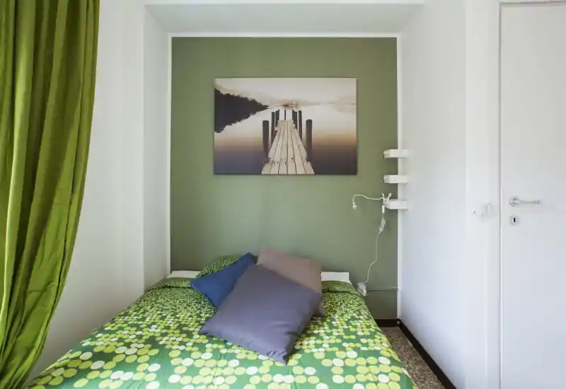 Chambre compacte et bien décorée avec un mur vert, une literie verte à motifs et des coussins décoratifs ; tableau au-dessus du lit et lumière naturelle douce créent une ambiance chaleureuse.