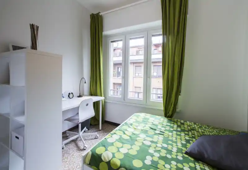 Chambre compacte et lumineuse avec un lit double à couette verte à motifs, un bureau blanc et une chaise près d'une grande fenêtre avec rideaux verts laissant entrer la lumière naturelle.