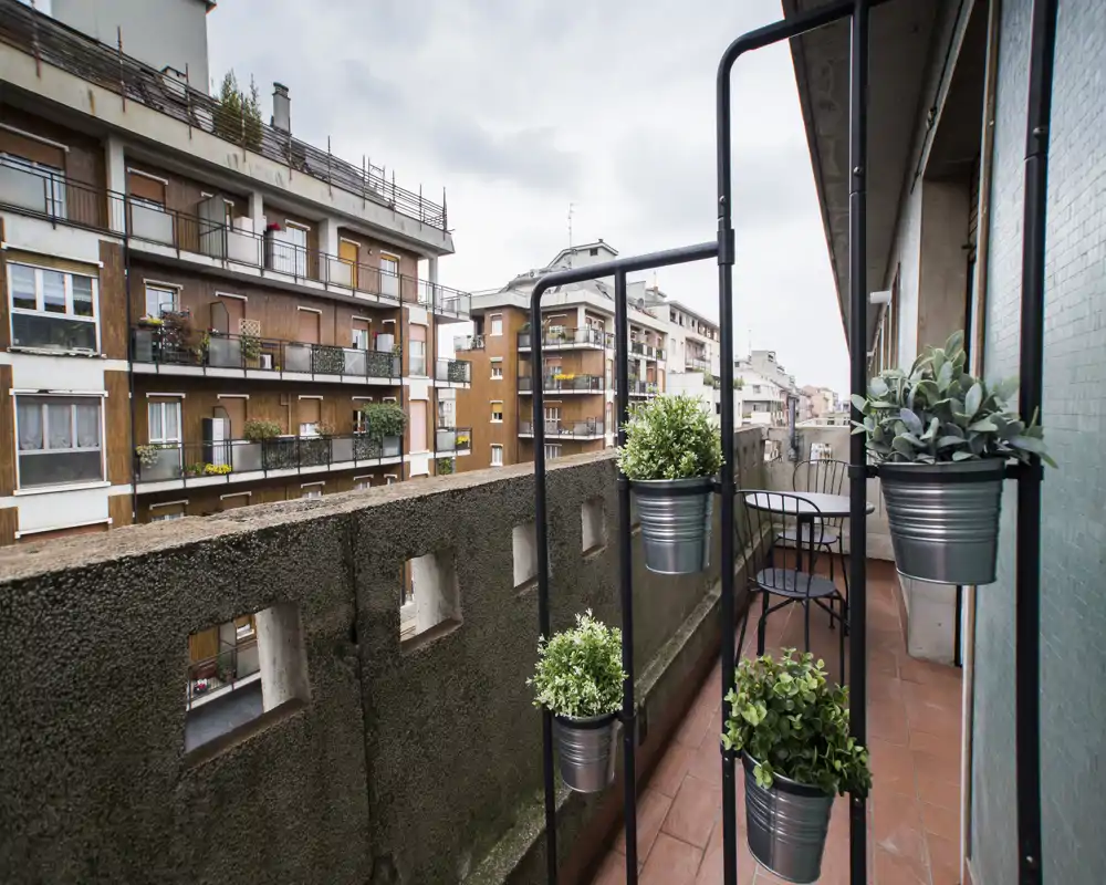 Sunny urban balcony with terracotta tile floor, metal bistro table and chairs, and vertical planters; overlooks neighboring apartment buildings.