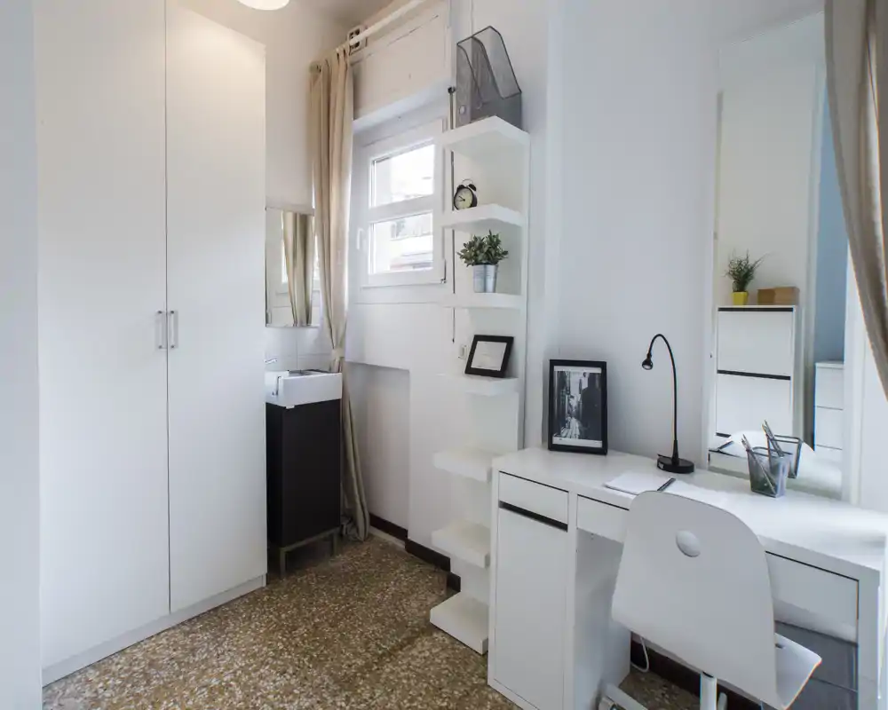 Compact, bright private room with white built-in wardrobe, shelving and a tidy desk — ideal for remote work or study. Natural light from the window keeps the space airy and pleasant.