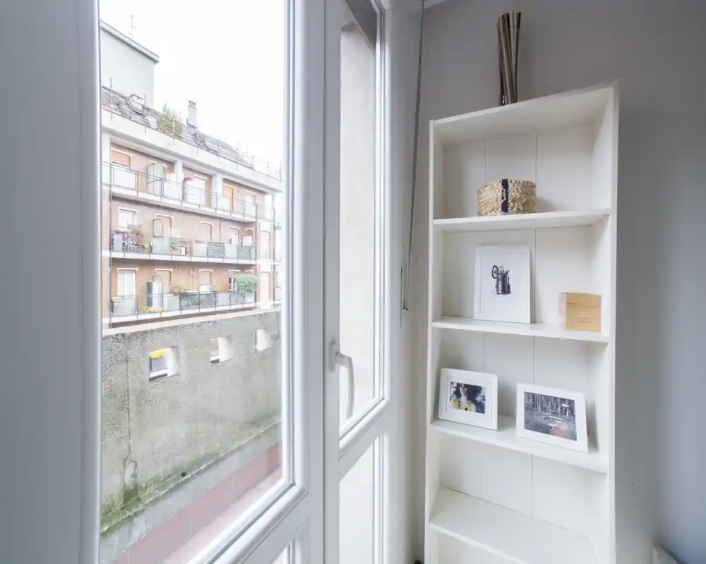 Bright corner featuring a tall white shelving unit beside a large window with a view onto adjacent balconies and a courtyard wall.