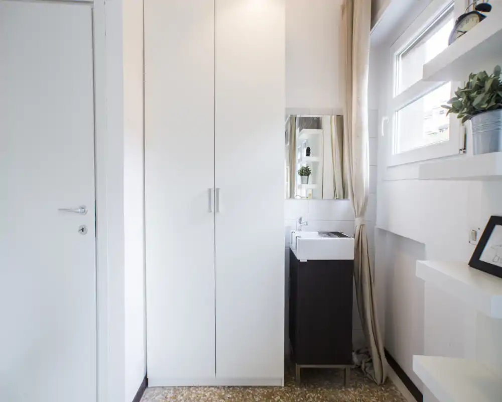 Compact bathroom corner with a small sink, mirror and storage cabinet; bright natural light from the window and neutral, modern decor.