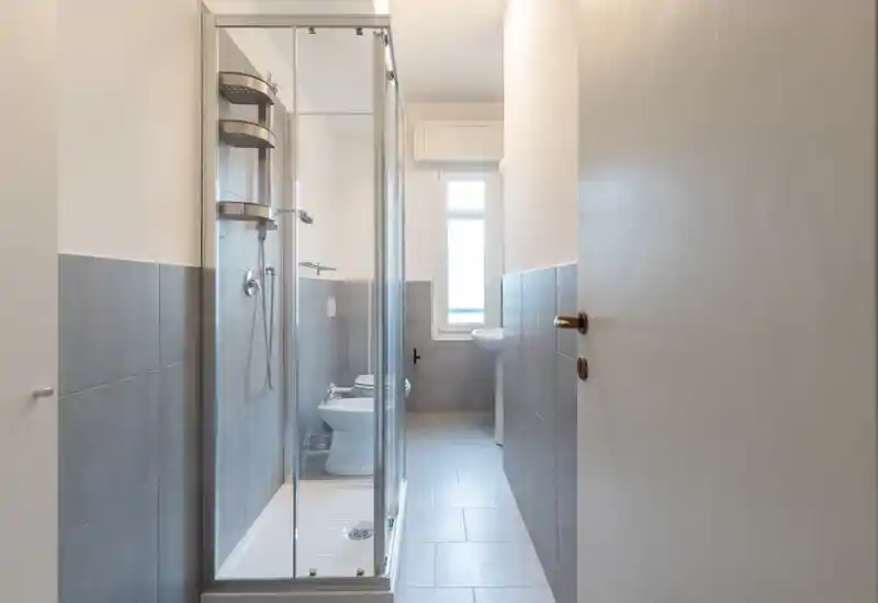 Compact modern bathroom with a glass-enclosed shower, wall-mounted fixtures, bidet and a pedestal sink. Neutral gray tiles and natural light from a narrow window create a clean, minimal look.