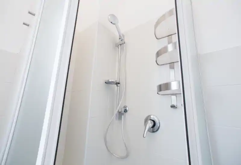 Bright, modern shower stall with a hand shower, chrome fixtures and wall-mounted shelving; clean white tiles and glass door.