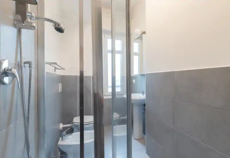 Bright, modern bathroom with a glass shower enclosure, wall-mounted fixtures, toilet and bidet visible, and a pedestal sink by the window.