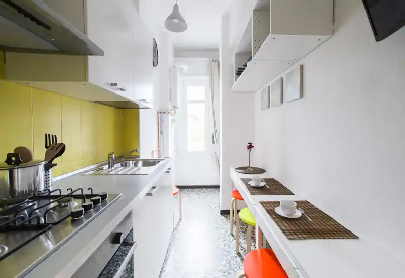Bright narrow kitchen with yellow tiled backsplash, stainless steel sink and gas hob; a slim breakfast counter with colorful stools and table settings makes this space functional and cheerful.
