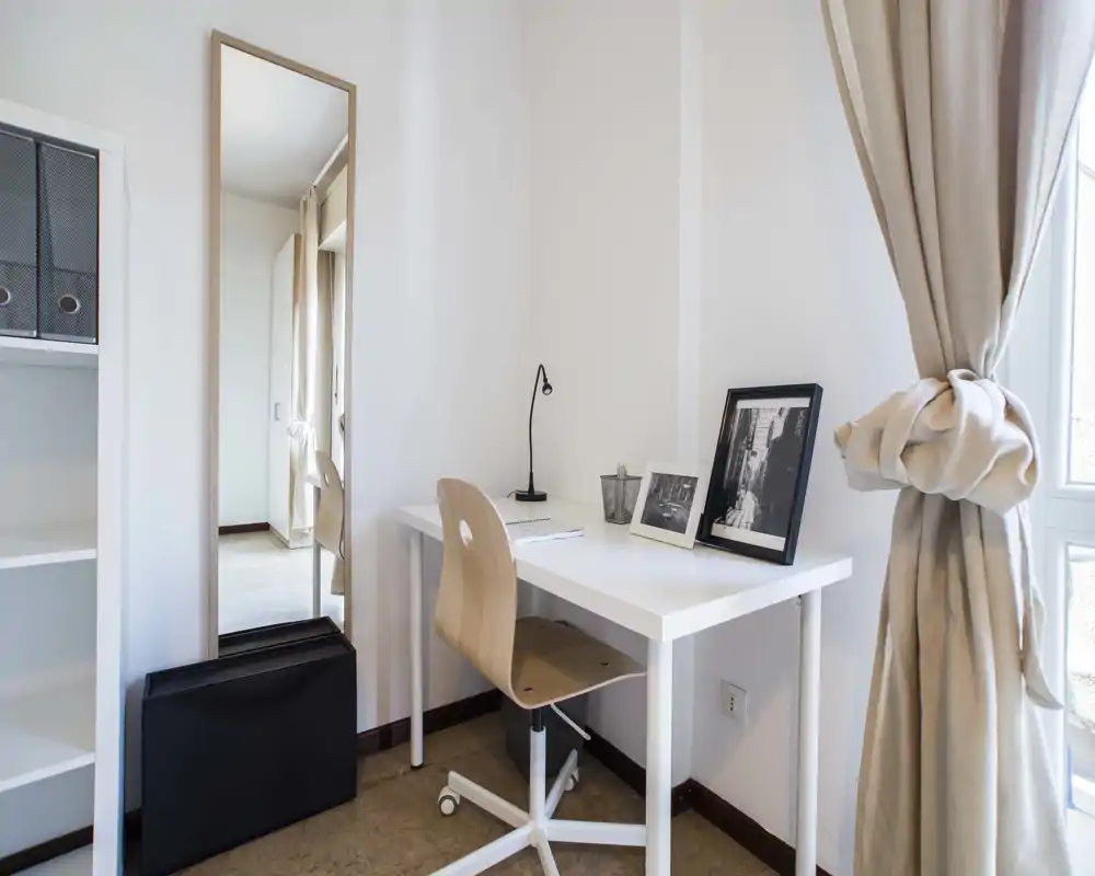 Espace de travail à domicile lumineux et minimaliste avec bureau blanc, chaise pivotante en bois et photos encadrées près d'une fenêtre aux rideaux beige.