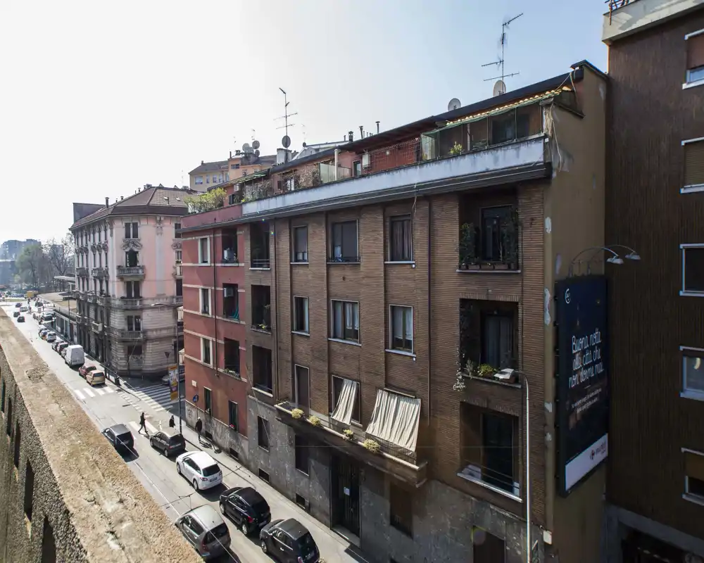 Vue de rue urbaine montrant une rangée d'immeubles résidentiels, des voitures stationnées et un trottoir ; éclairage de jour clair et détails architecturaux visibles.