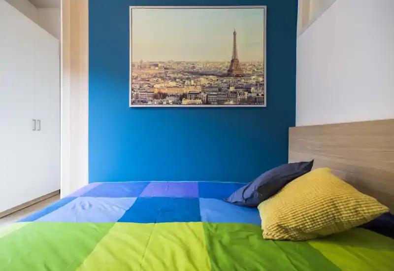 Chambre lumineuse et colorée avec un grand lit habillé d'une couette bleu et verte, une tête de lit en bois et des coussins décoratifs ; un tableau de paysage urbain est accroché sur un mur bleu profond.