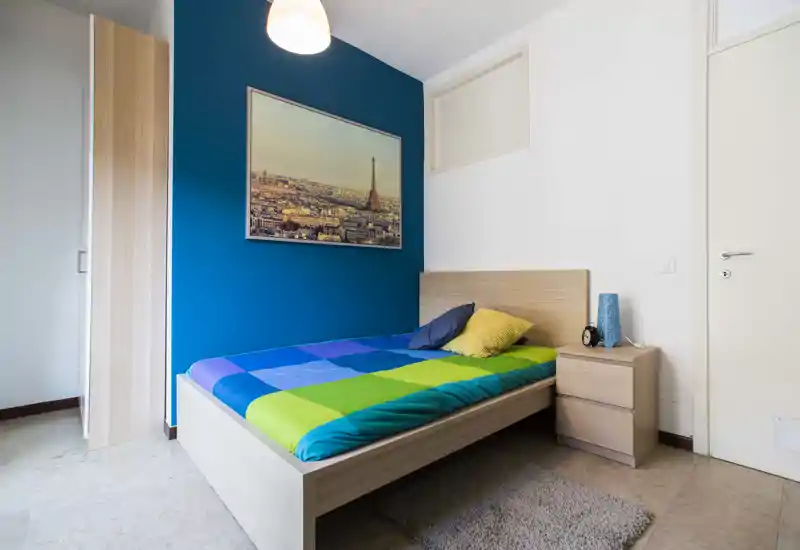 Chambre simple soignée avec lit coloré, mur d'accent bleu et table de chevet. Espace lumineux et bien aménagé, parfait pour une personne seule.
