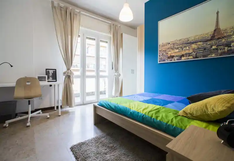 Chambre moderne et lumineuse avec un lit bas en bois, une literie colorée et un mur d'accent bleu. Grande fenêtre avec voilages laissant entrer la lumière naturelle; bureau et placard compacts pour un usage pratique.