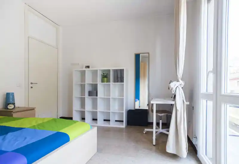 Chambre moderne et lumineuse avec un lit coloré au premier plan, une étagère blanche, un bureau près de la fenêtre et une grande glace pleine longueur; de larges fenêtres apportent beaucoup de lumière naturelle.